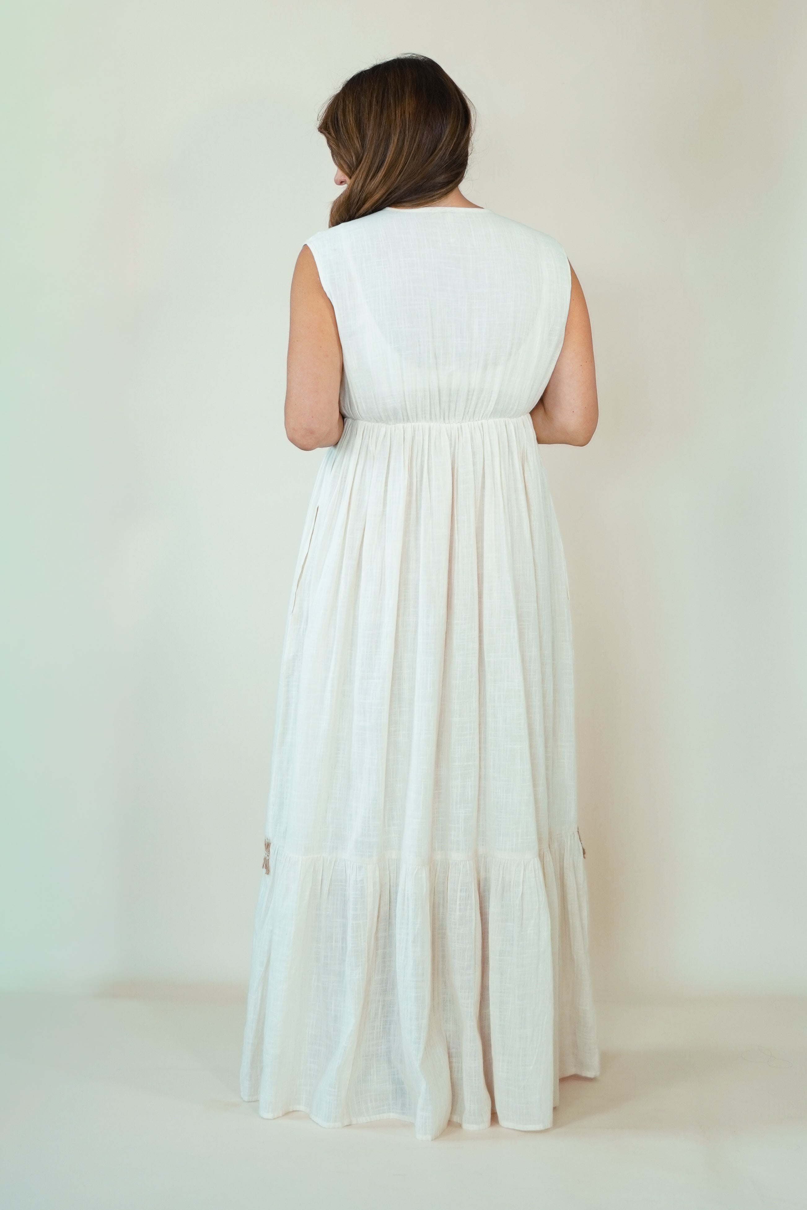 A woman viewed from behind wearing a sleeveless, tiered white maxi dress against a plain, warm off-white studio background.