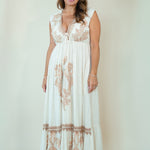 A woman modeling a sleeveless white maxi dress with beige floral embroidery and a deep V-neckline, standing against a plain light beige studio background.