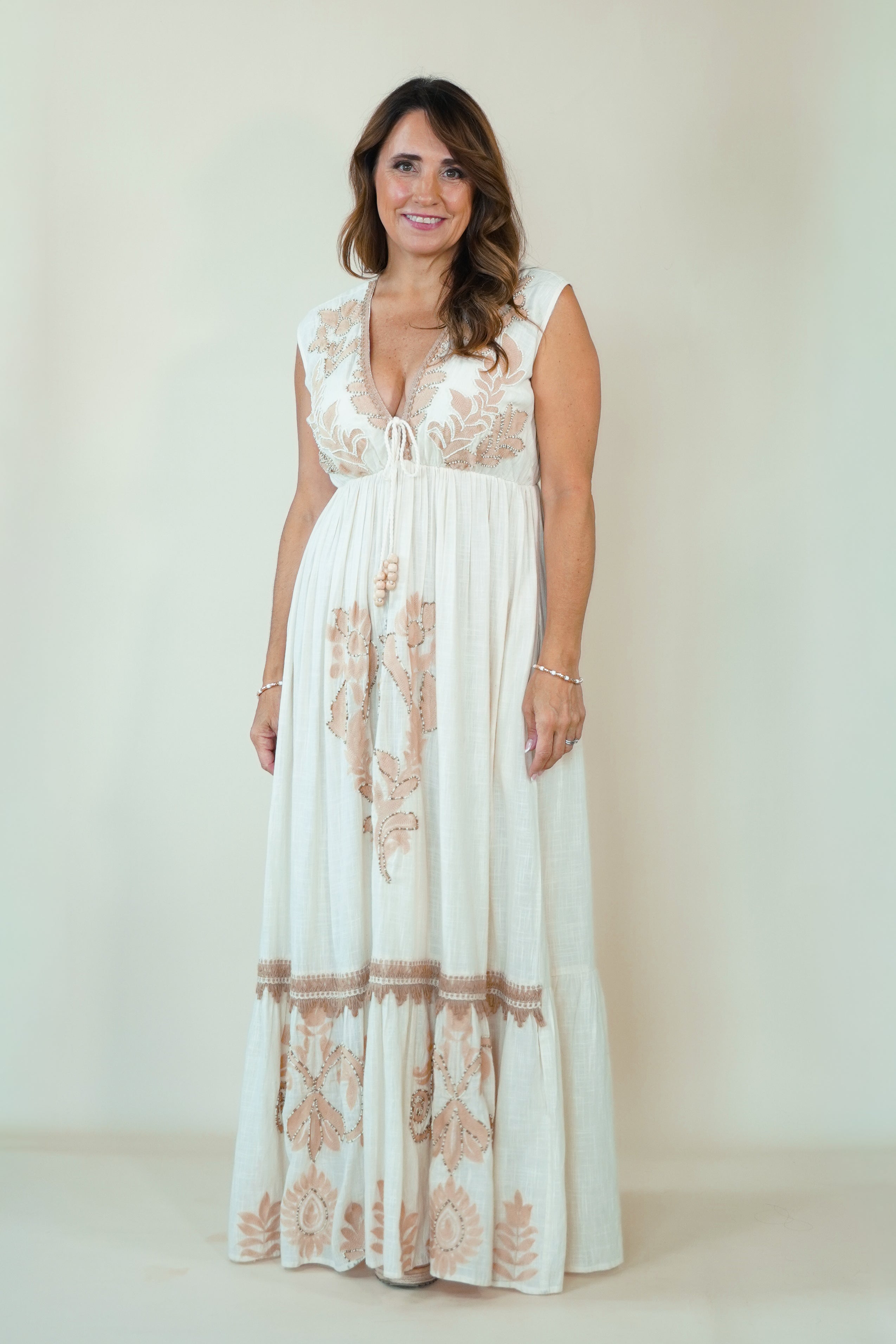 A woman modeling a sleeveless white maxi dress with beige floral embroidery and a deep V-neckline, standing against a plain light beige studio background.