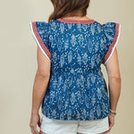 A woman shown from the back wearing a blue patterned short-sleeve top with red and white trim, white shorts, and tan wedge sandals against a plain beige studio background.