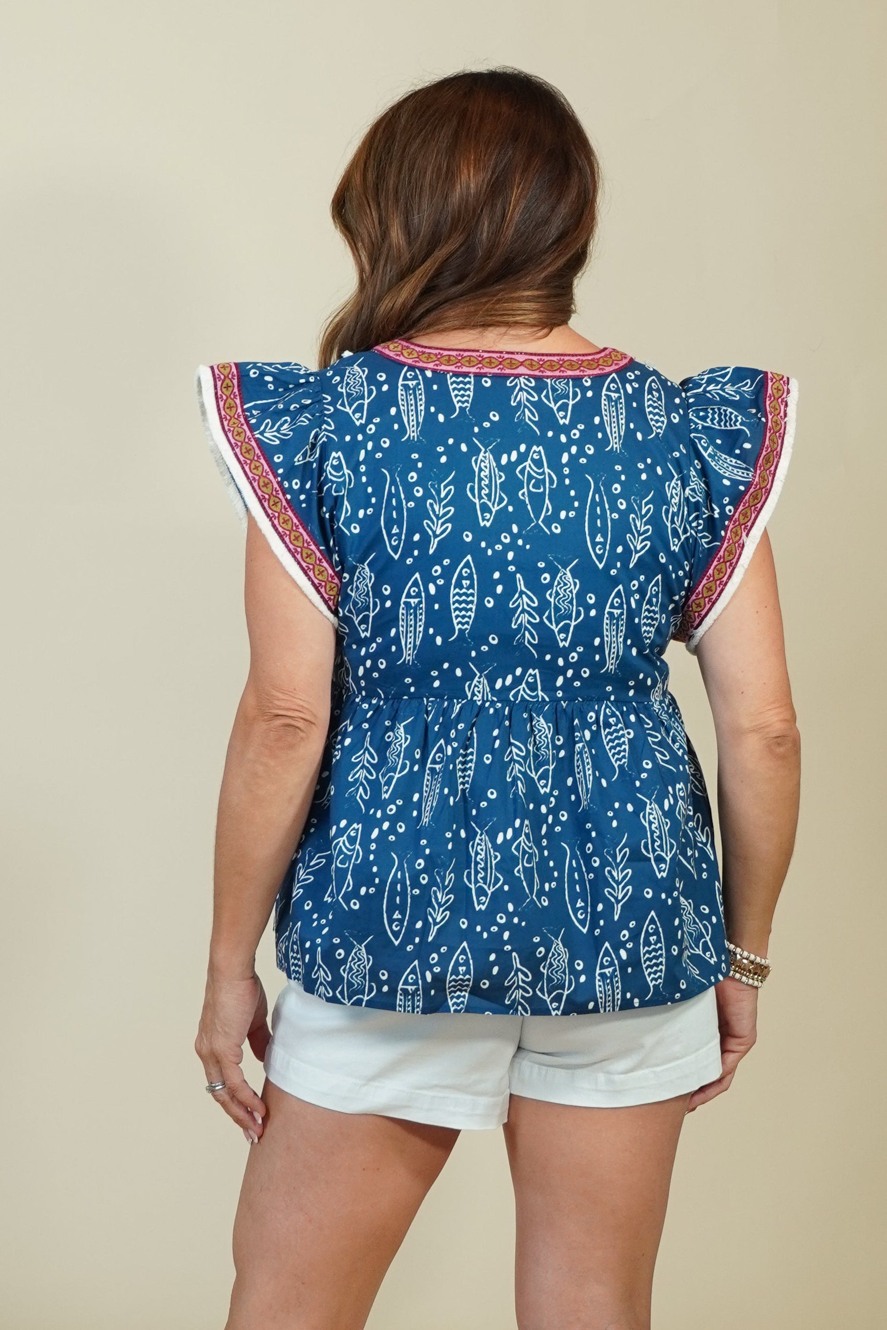 A woman shown from the back wearing a blue patterned short-sleeve top with red and white trim, white shorts, and tan wedge sandals against a plain beige studio background.