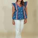 A woman modeling a blue patterned peplum top with red and white striped trim, paired with white pants, against a solid beige studio background.