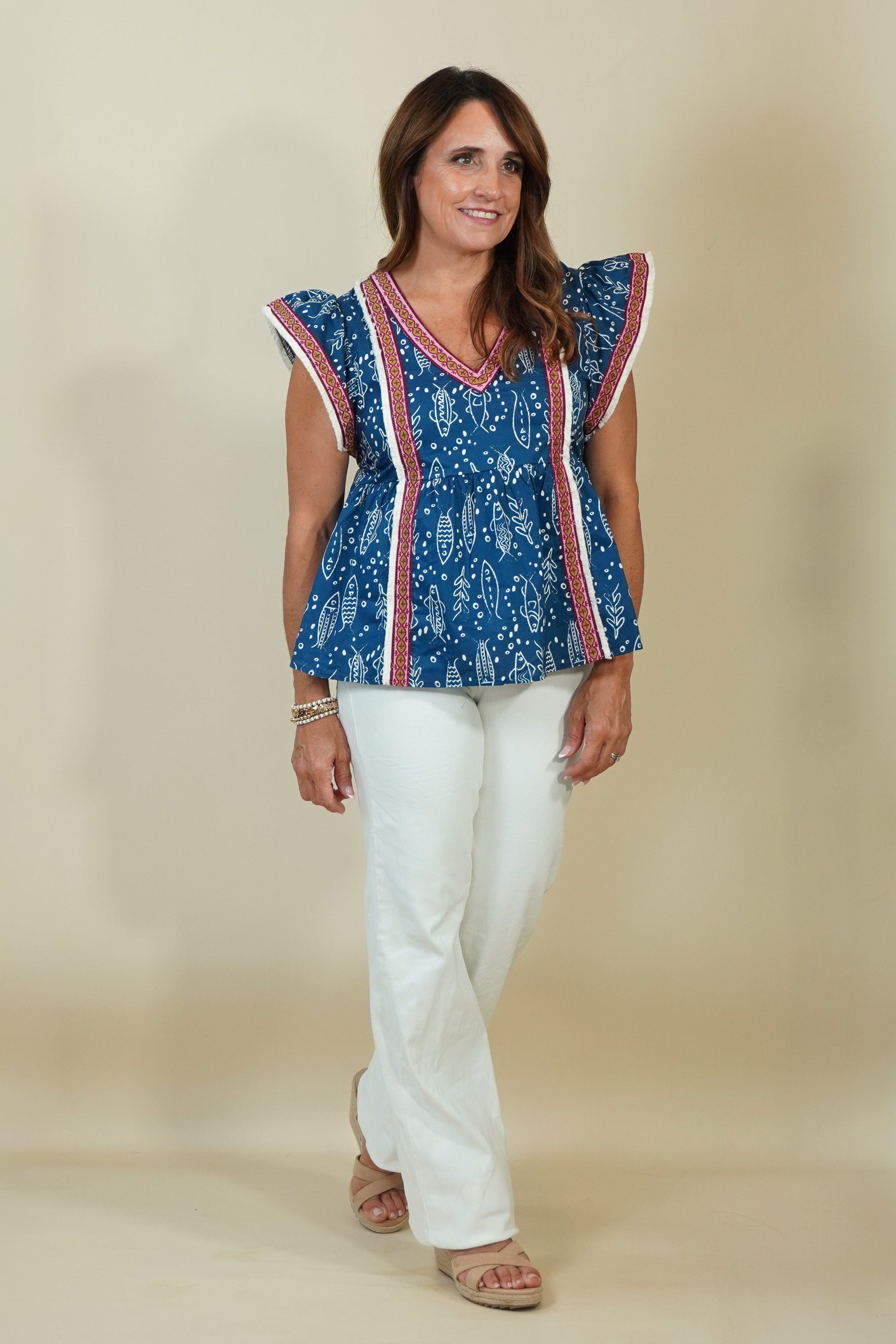 A woman modeling a blue patterned peplum top with red and white striped trim, paired with white pants, against a solid beige studio background.