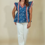 A woman modeling a blue patterned short-sleeve blouse with red and white striped trim, paired with white pants, standing against a plain beige studio backdrop.