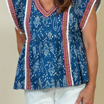 A woman modeling a blue patterned short-sleeve top with red and white striped trim, paired with white shorts and beige sandals, standing against a plain beige studio background.