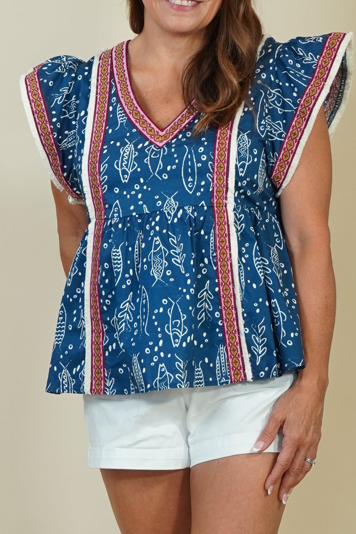 A woman modeling a blue patterned short-sleeve top with red and white striped trim, paired with white shorts and beige sandals, standing against a plain beige studio background.