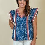 A woman modeling a sleeveless blue patterned top with red and white trim, paired with white shorts and beige wedge sandals, against a plain beige studio background.