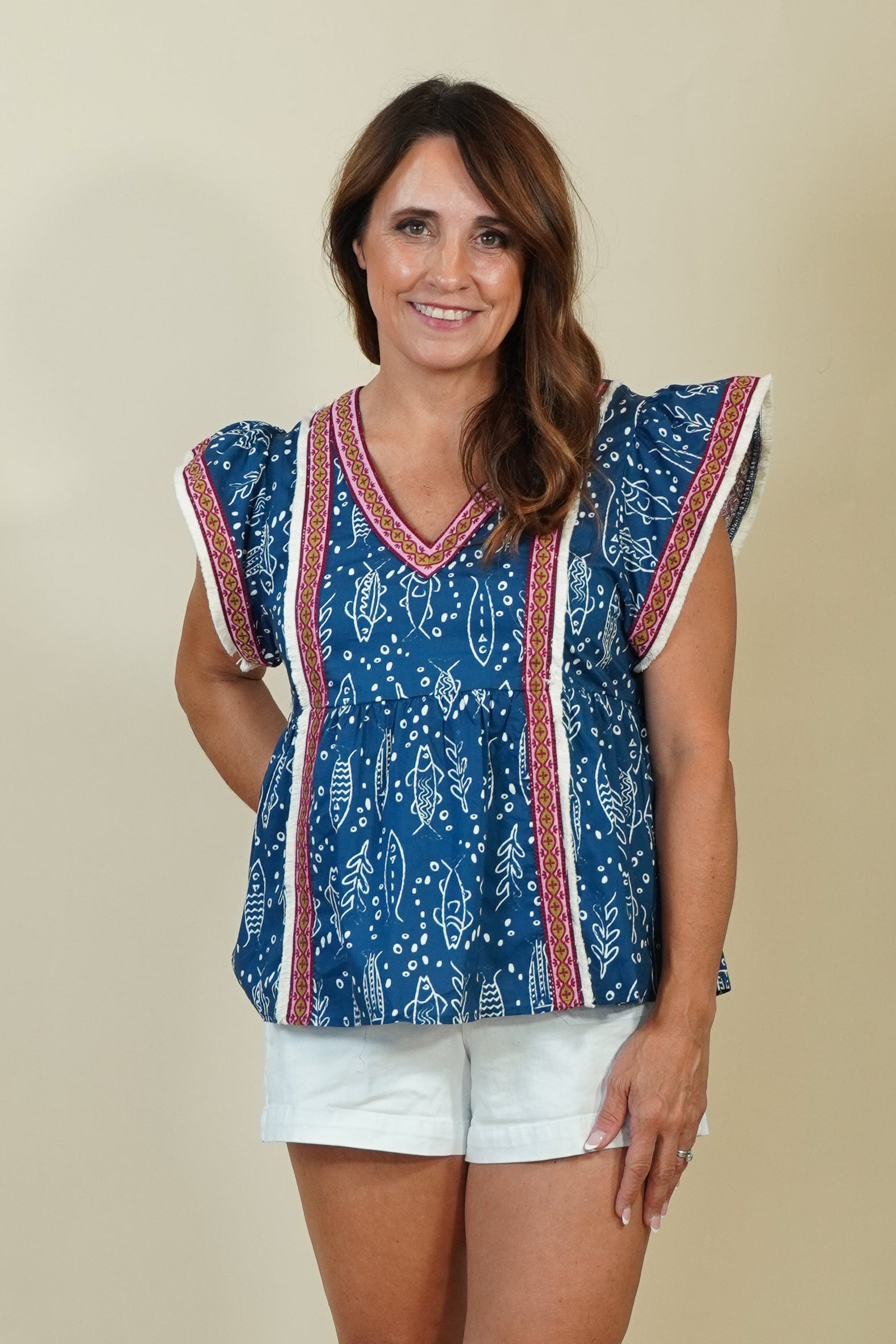 A woman modeling a sleeveless blue patterned top with red and white trim, paired with white shorts and beige wedge sandals, against a plain beige studio background.