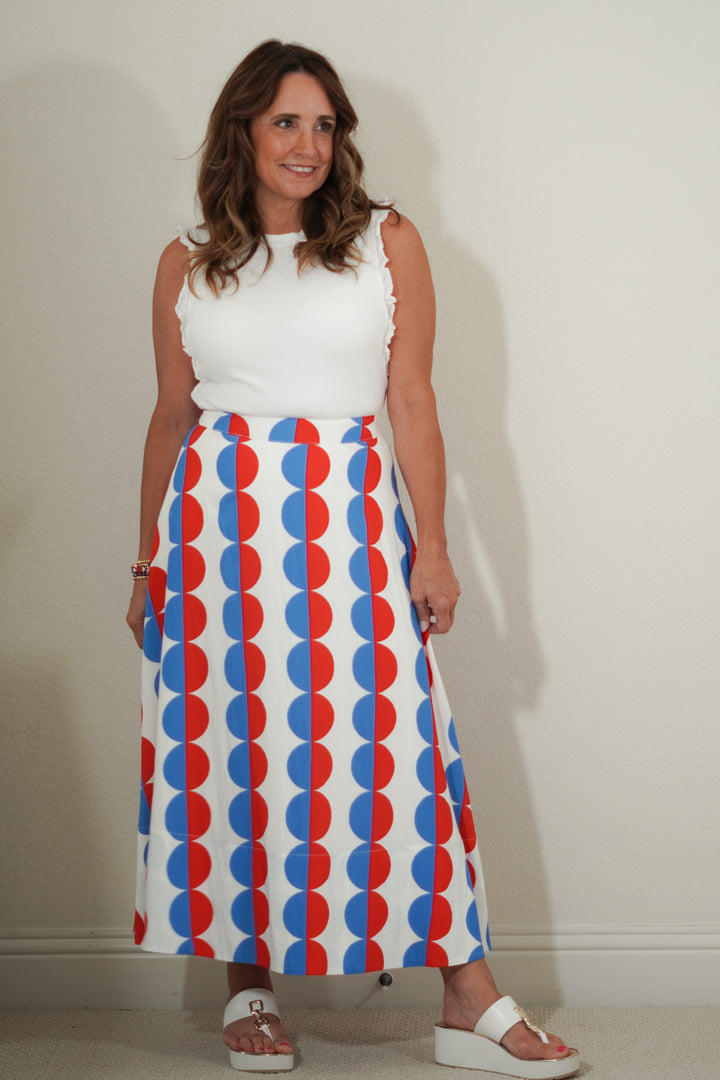 Woman wearing a white sleeveless top and a colorful skirt with red, blue, and white pattern against a plain background.