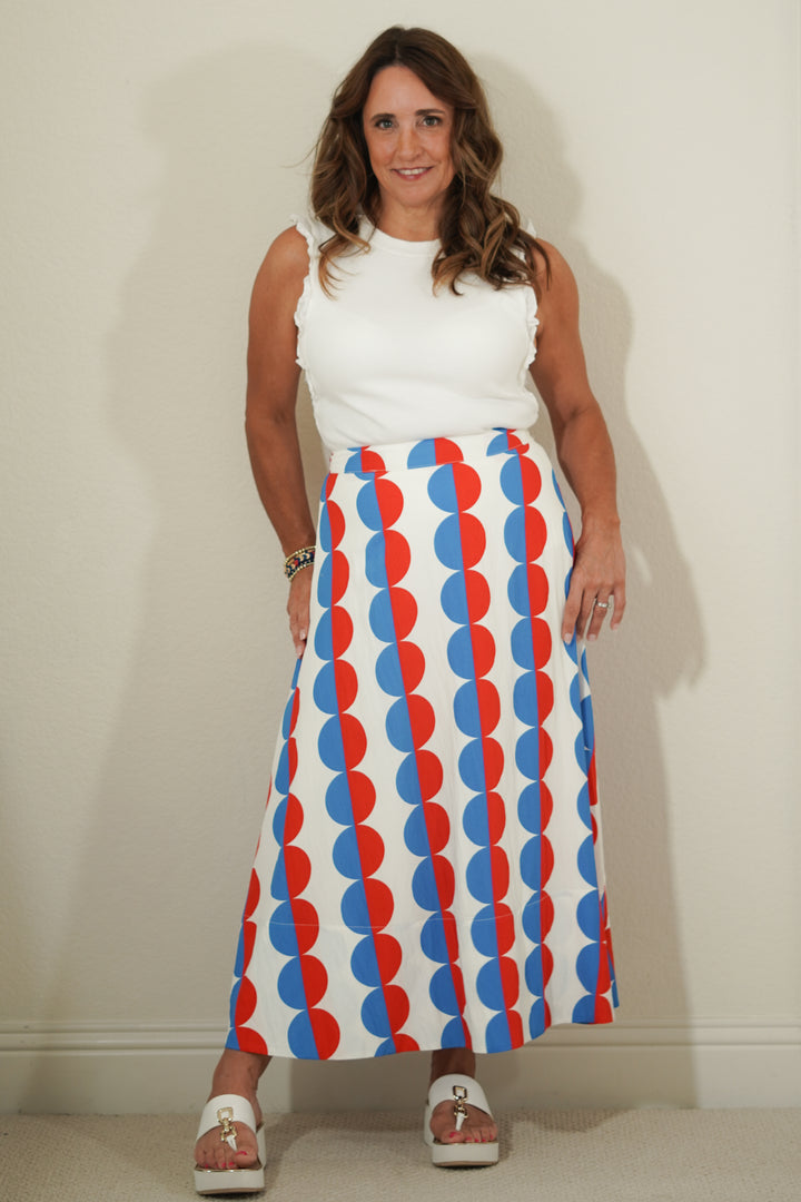 Woman wearing a white sleeveless top and a long, patterned skirt with red and blue circles on a plain background.