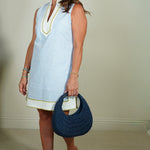 Woman wearing a light blue sleeveless dress with a white collar, holding a navy blue handbag indoors.