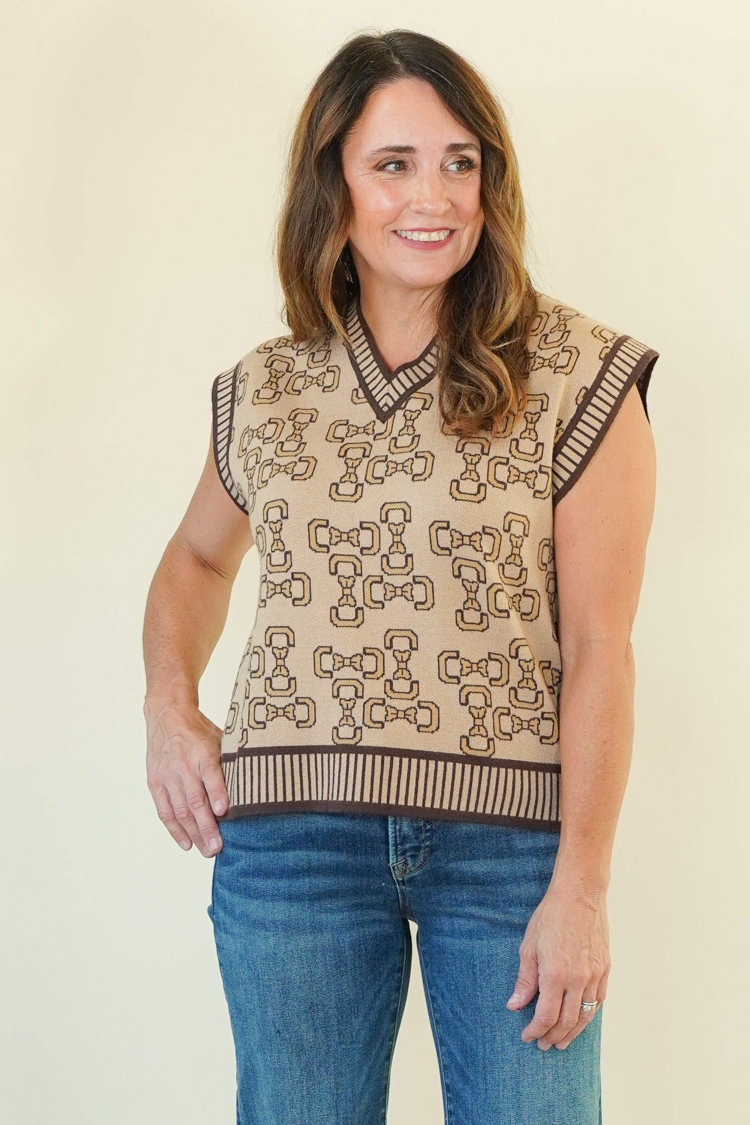 A woman modeling a sleeveless knit vest with a beige and brown geometric pattern, paired with blue jeans, standing against a plain cream background.