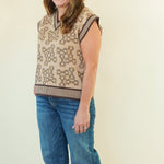A woman modeling a sleeveless knit sweater vest with a beige and brown geometric pattern, paired with blue jeans and brown shoes, standing against a plain cream background.