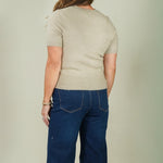 A woman shown from the back wearing a light beige short-sleeve knit top and dark blue wide-leg jeans, standing against a plain light gray studio background.