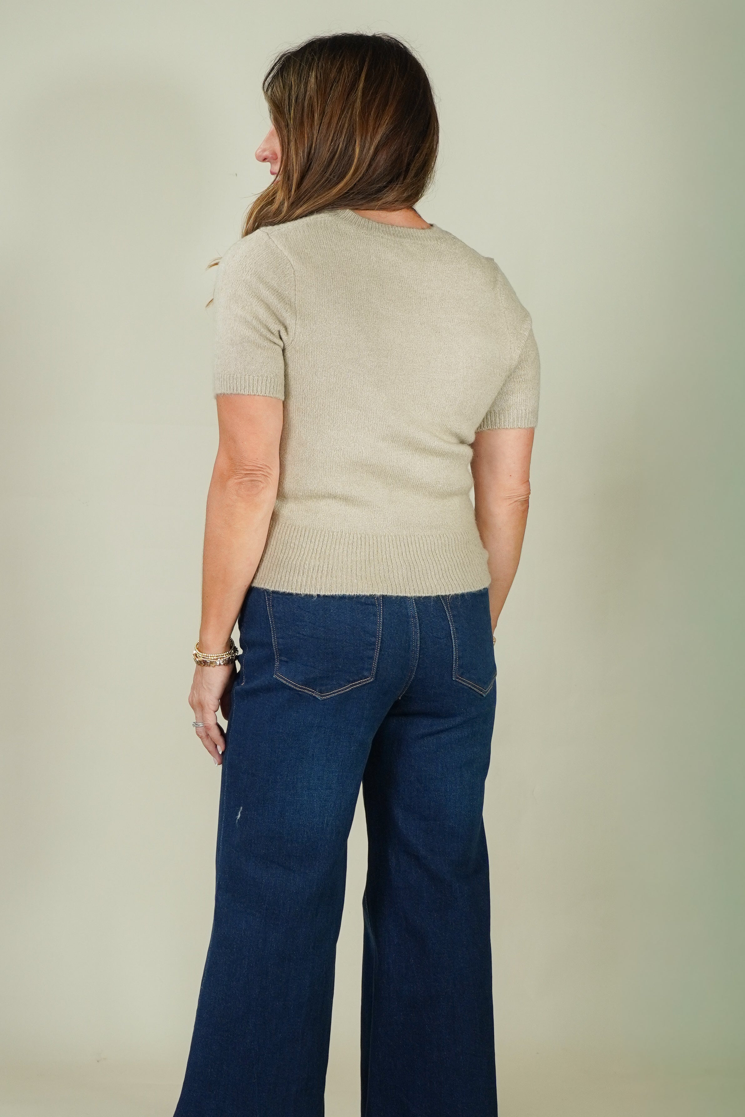 A woman shown from the back wearing a light beige short-sleeve knit top and dark blue wide-leg jeans, standing against a plain light gray studio background.