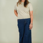 A woman modeling a beige short-sleeve knit top and dark blue wide-leg jeans against a plain light beige studio background.