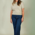 A woman modeling a beige short-sleeve knit top and dark blue wide-leg jeans against a plain light beige studio backdrop.