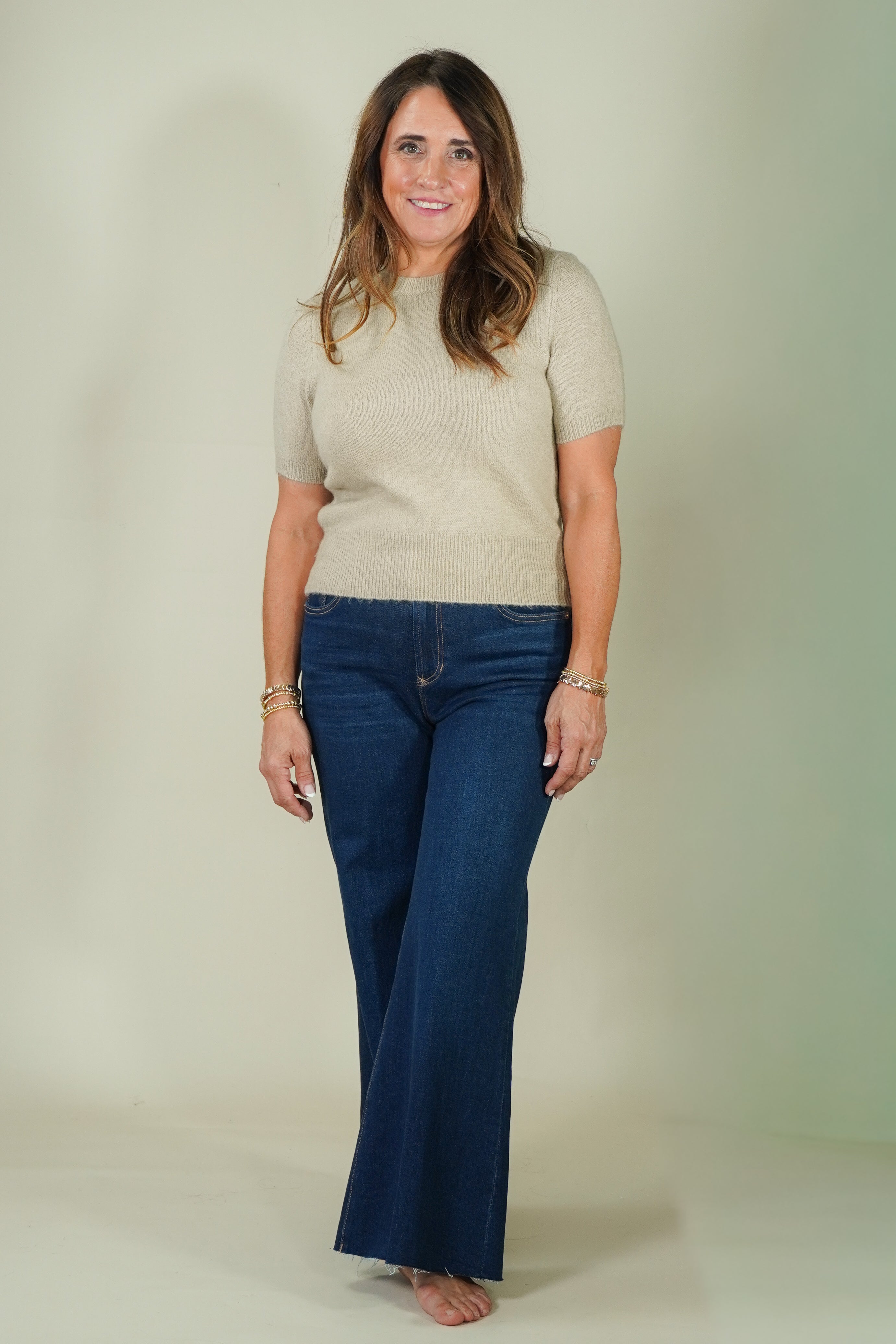 A woman modeling a beige short-sleeve knit top and dark blue wide-leg jeans against a plain light beige studio backdrop.