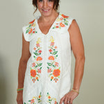 Woman wearing a white dress with floral embroidery against a plain background