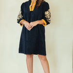 A woman modeling a dark navy blue tunic dress with yellow trim at the collar and cuffs, and gold floral embroidery on the sleeves, paired with beige wedge sandals against a plain cream background.