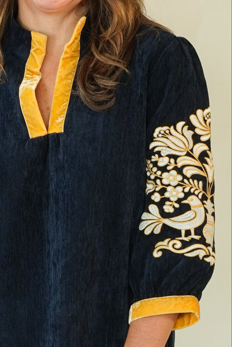 A woman modeling a dark navy blue tunic dress with yellow trim and gold floral embroidery on the sleeves, paired with beige wedge sandals, against a plain cream background.