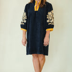 A woman modeling a dark navy blue tunic dress with gold embroidery on the sleeves and a contrasting gold collar, paired with beige wedge sandals against a plain cream background.