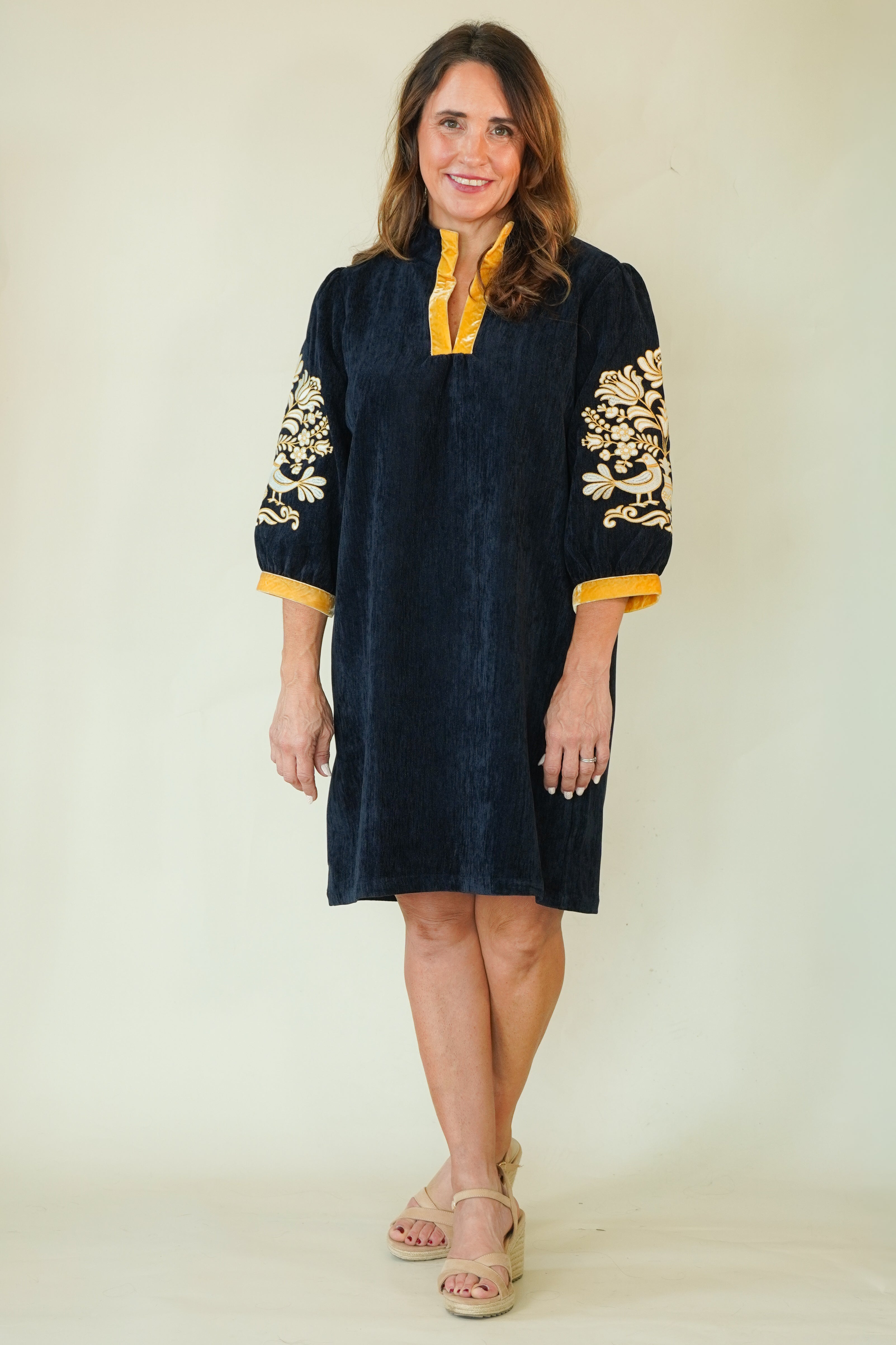 A woman modeling a dark navy blue tunic dress with gold embroidery on the sleeves and a contrasting gold collar, paired with beige wedge sandals against a plain cream background.