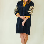 A woman modeling a dark navy blue velvet tunic dress with yellow trim and gold floral embroidery on the sleeves, paired with beige wedge sandals, against a plain cream background.