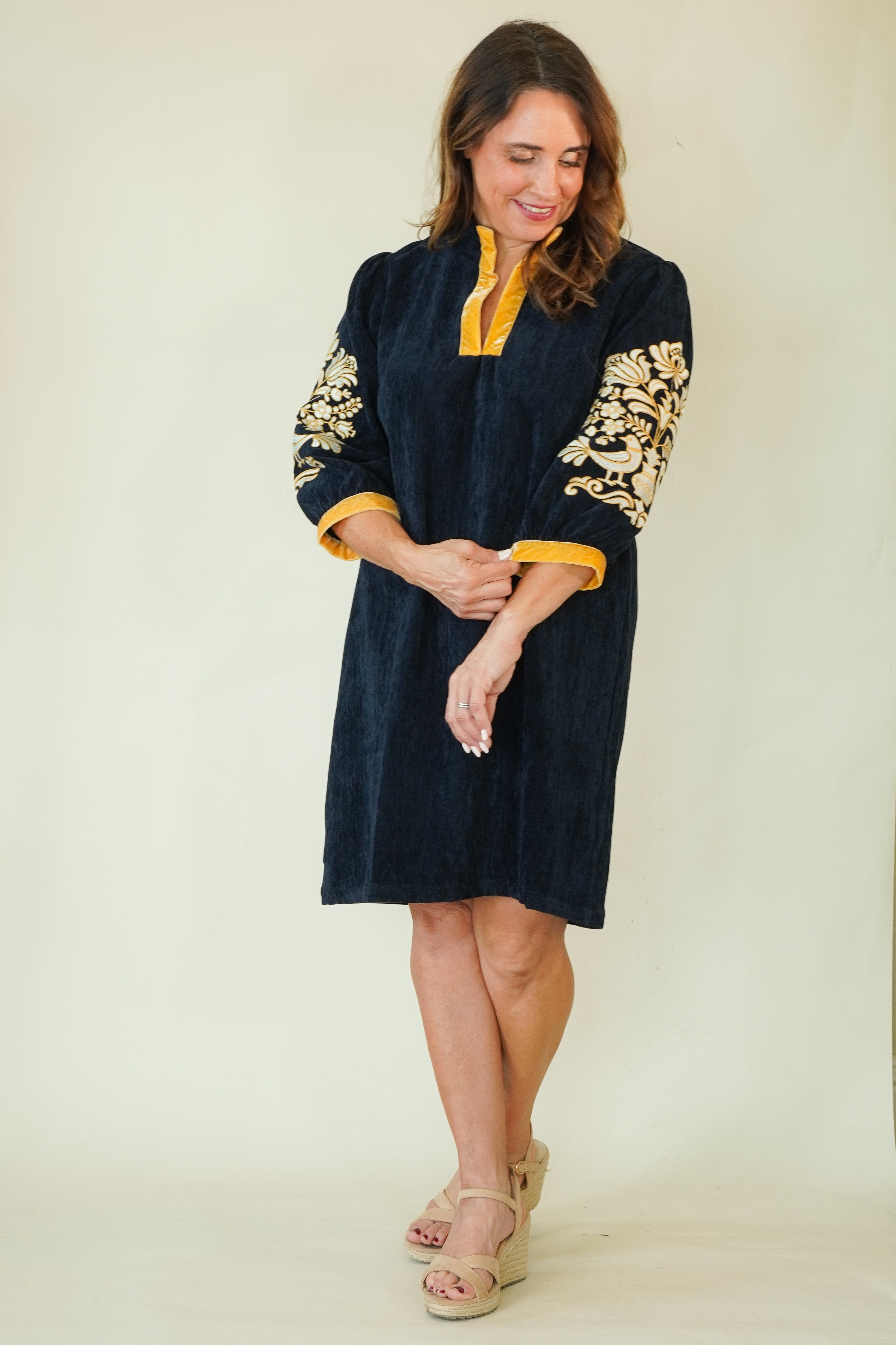 A woman modeling a dark navy blue velvet tunic dress with yellow trim and gold floral embroidery on the sleeves, paired with beige wedge sandals, against a plain cream background.