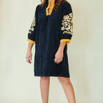A woman modeling a dark navy blue tunic dress with gold floral embroidery on the sleeves and a yellow collar, paired with beige wedge sandals against a plain cream background.