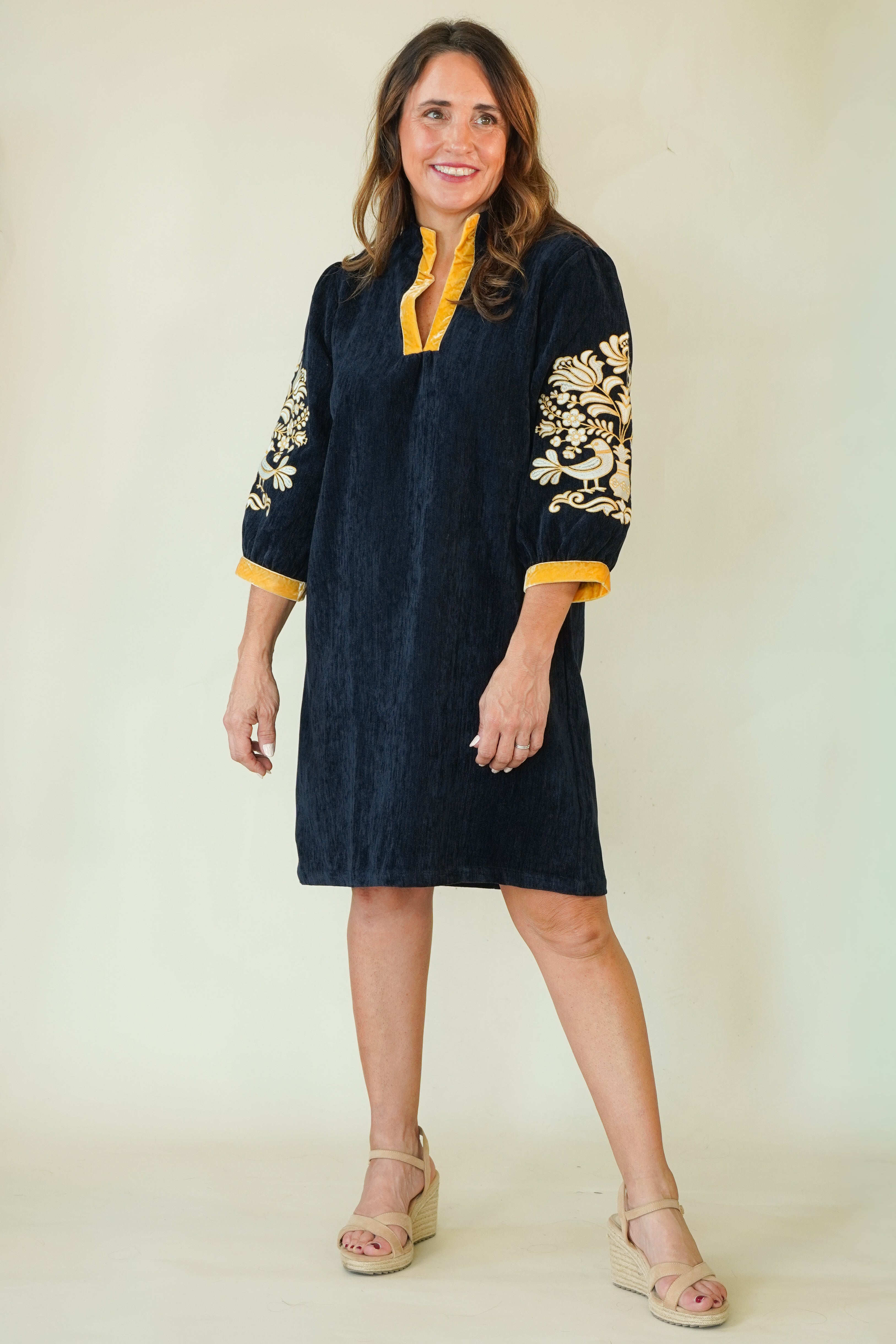 A woman modeling a dark navy blue tunic dress with gold floral embroidery on the sleeves and a yellow collar, paired with beige wedge sandals against a plain cream background.