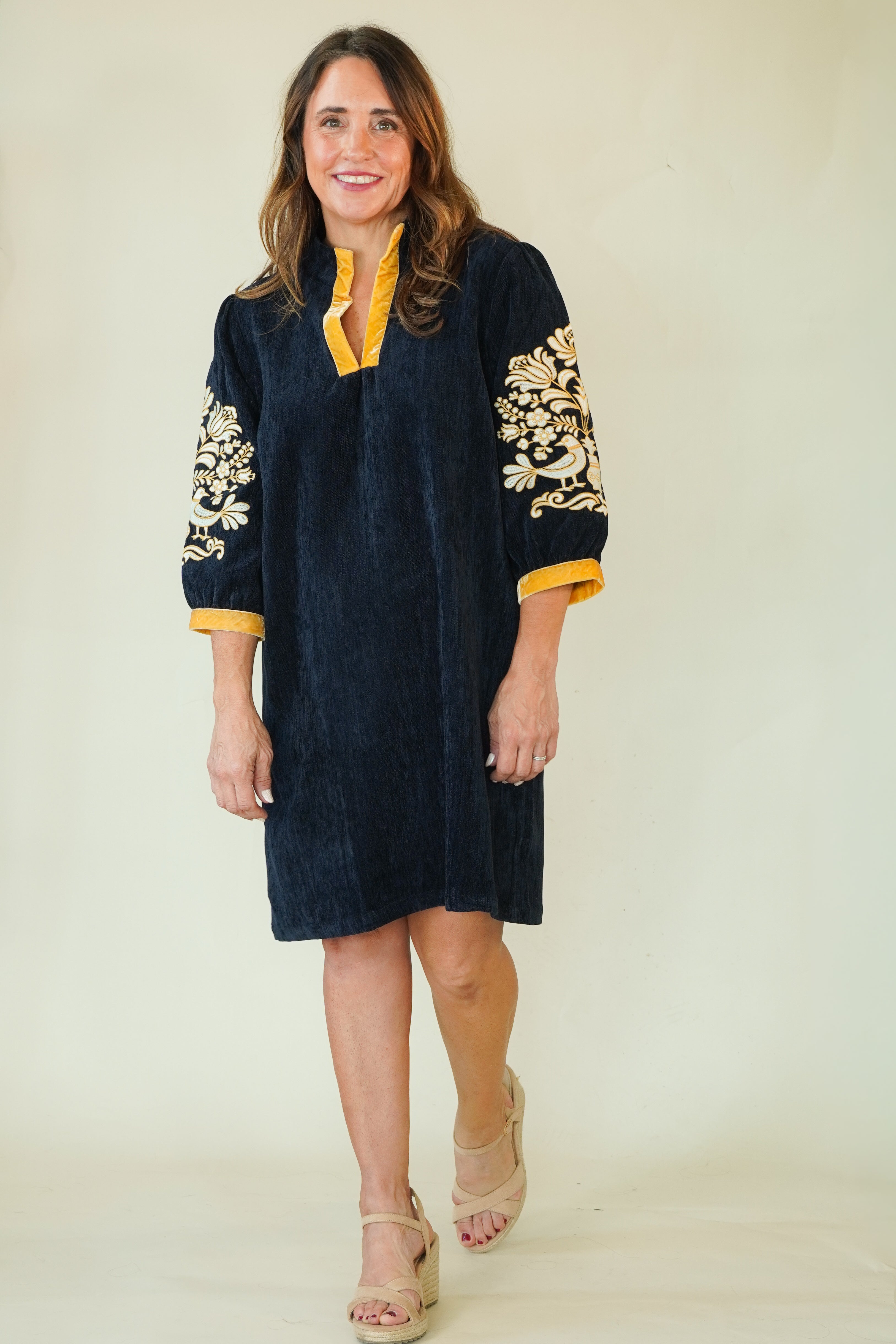 A woman modeling a dark navy blue tunic dress with yellow trim and gold floral embroidery on the sleeves, paired with beige wedge sandals, against a plain cream background.