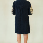 A woman viewed from the back wearing a dark navy corduroy tunic dress with gold floral embroidery on the sleeves, paired with beige espadrille wedge sandals, against a plain cream background.