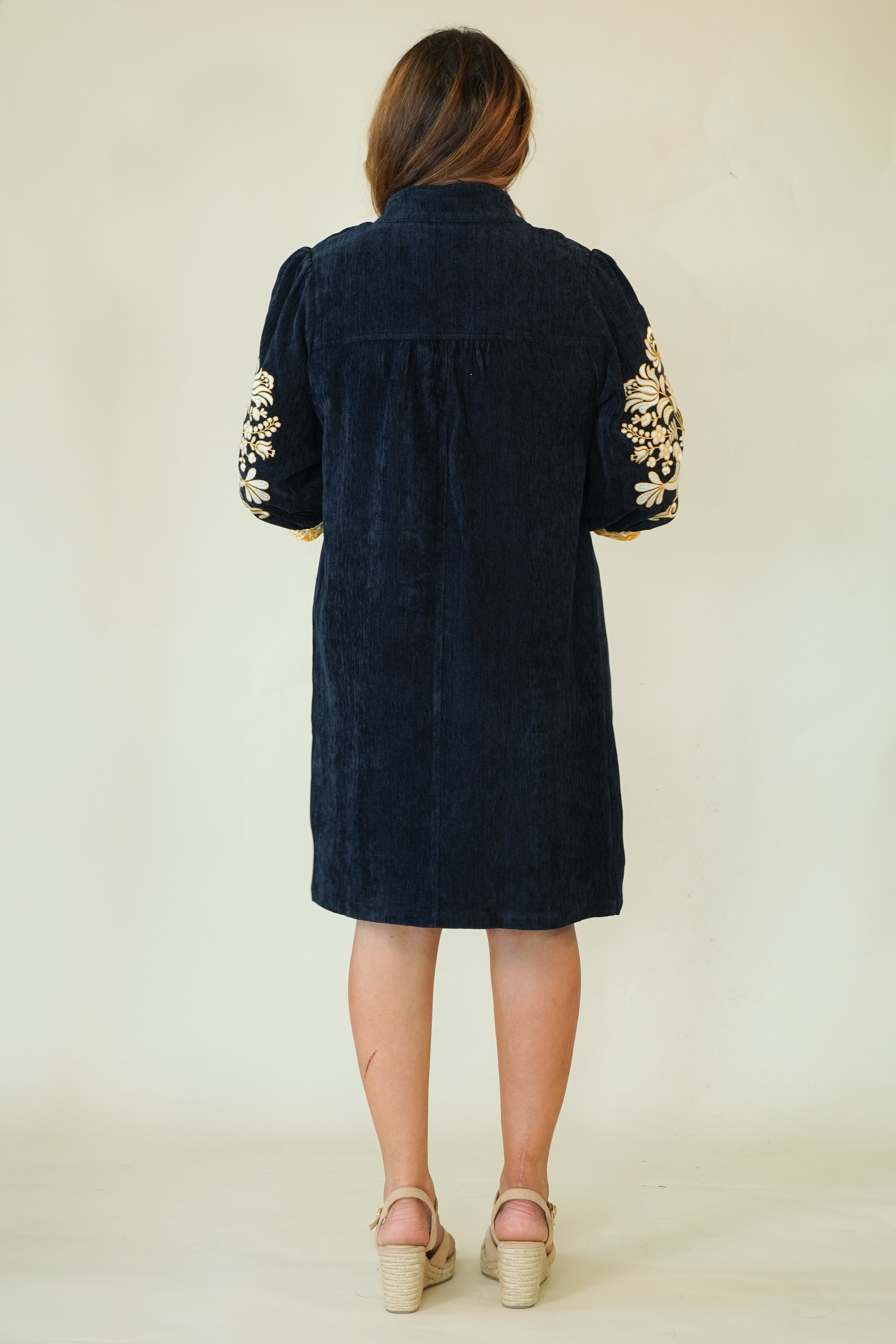 A woman viewed from the back wearing a dark navy corduroy tunic dress with gold floral embroidery on the sleeves, paired with beige espadrille wedge sandals, against a plain cream background.