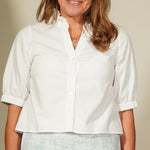 Woman wearing a white blouse and light blue shorts standing against a plain wall.