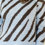 A woman modeling a light blue and dark brown zebra-striped knit sweater, paired with white pants and brown woven loafers, standing against a plain cream background.