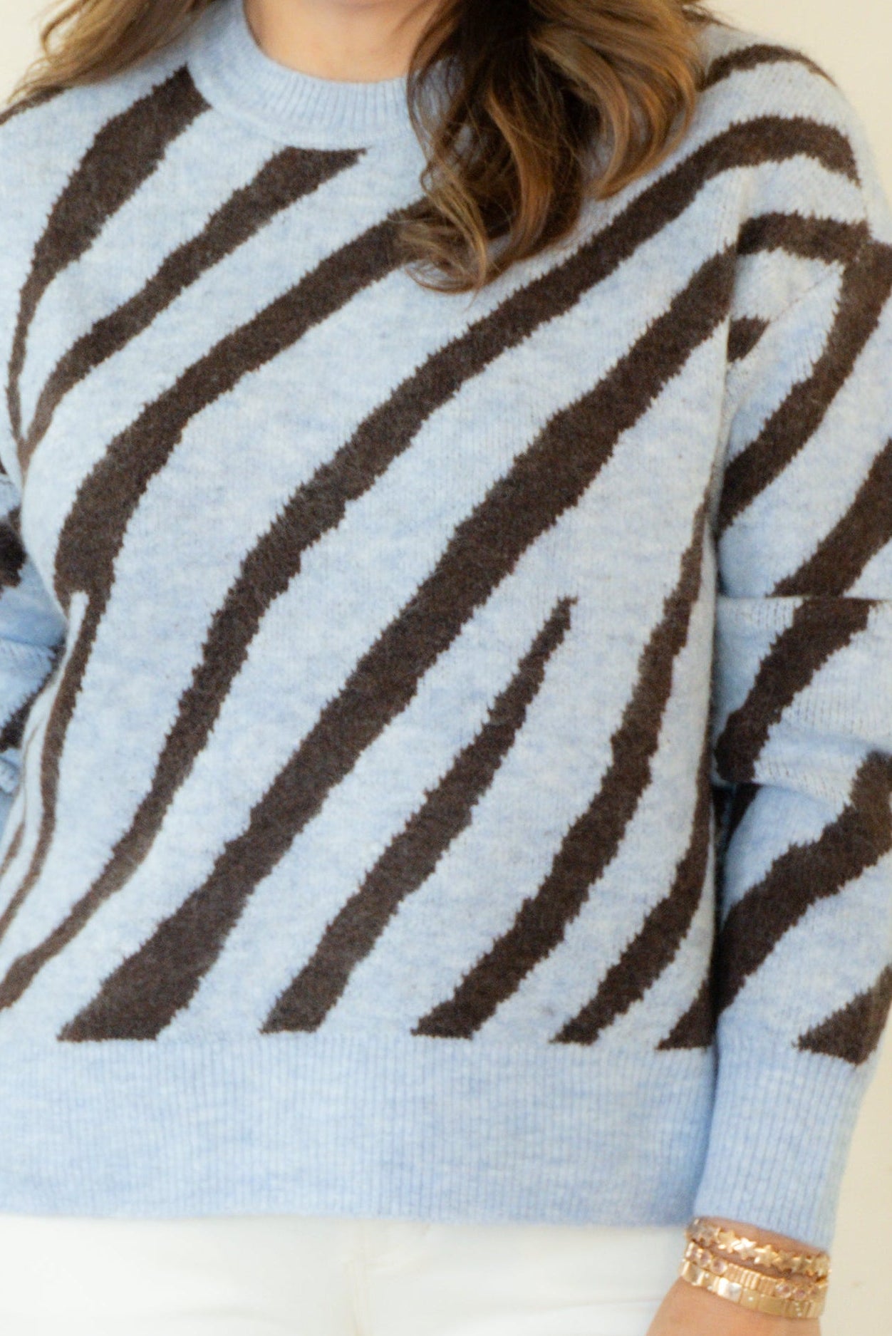 A woman modeling a light blue and dark brown zebra-striped knit sweater, paired with white pants and brown woven loafers, standing against a plain cream background.