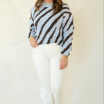 A woman modeling a light blue and dark brown zebra-striped knit sweater, paired with white pants and brown moccasin-style shoes, against a plain cream studio backdrop.