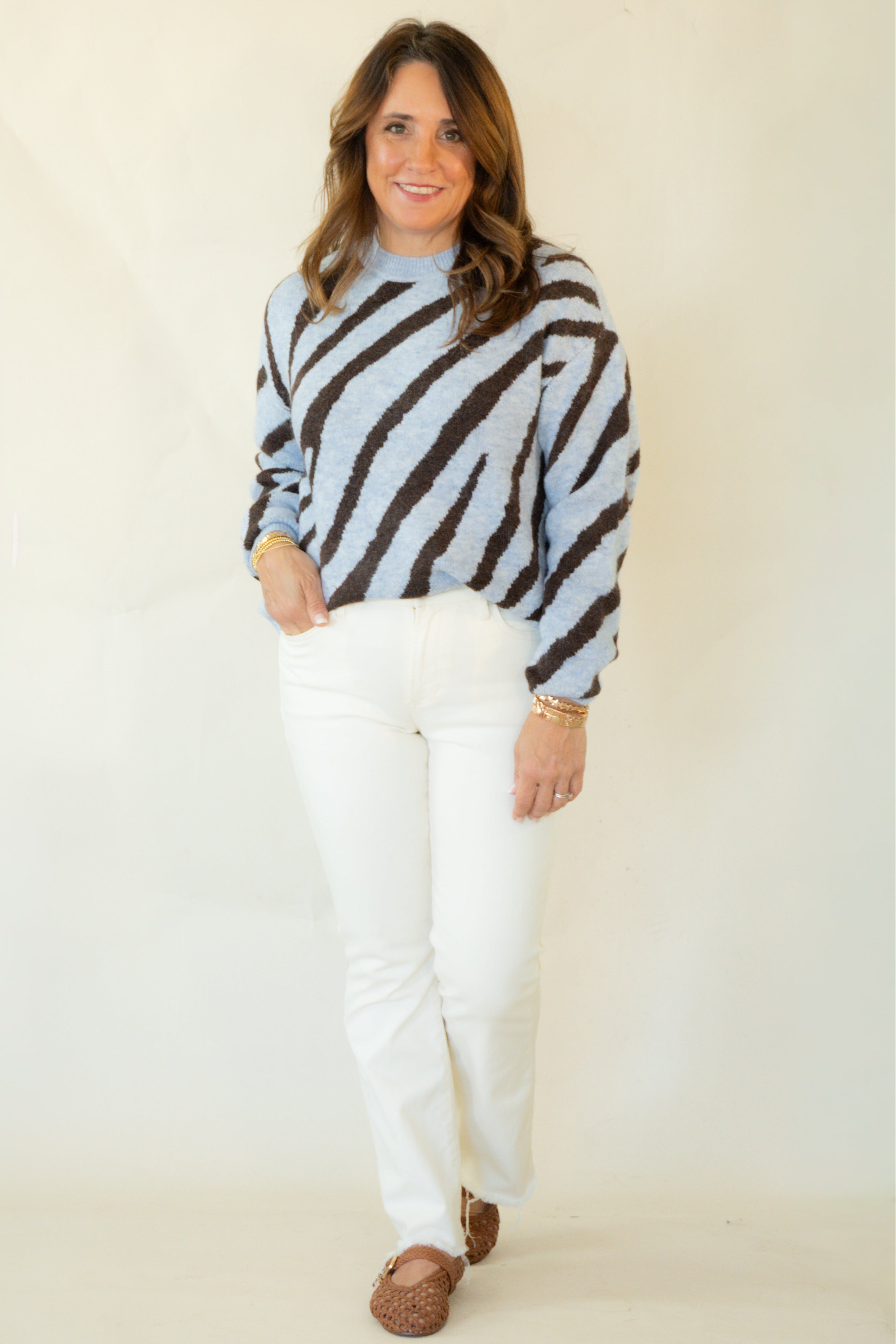 A woman modeling a light blue and dark brown zebra-striped knit sweater, paired with white pants and brown moccasin-style shoes, against a plain cream studio backdrop.
