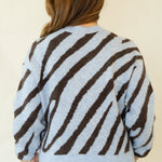 A woman viewed from behind wearing a light blue knit sweater with bold dark brown diagonal stripes, paired with white pants and brown lace-up sandals, against a plain cream background.