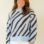 A woman modeling a light blue and dark brown striped knit sweater, paired with white pants and brown woven loafers, standing against a plain cream-colored studio backdrop.