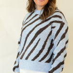 A woman modeling a light blue and dark brown zebra-striped knit sweater, paired with white pants and brown shoes, standing against a plain off-white studio background.