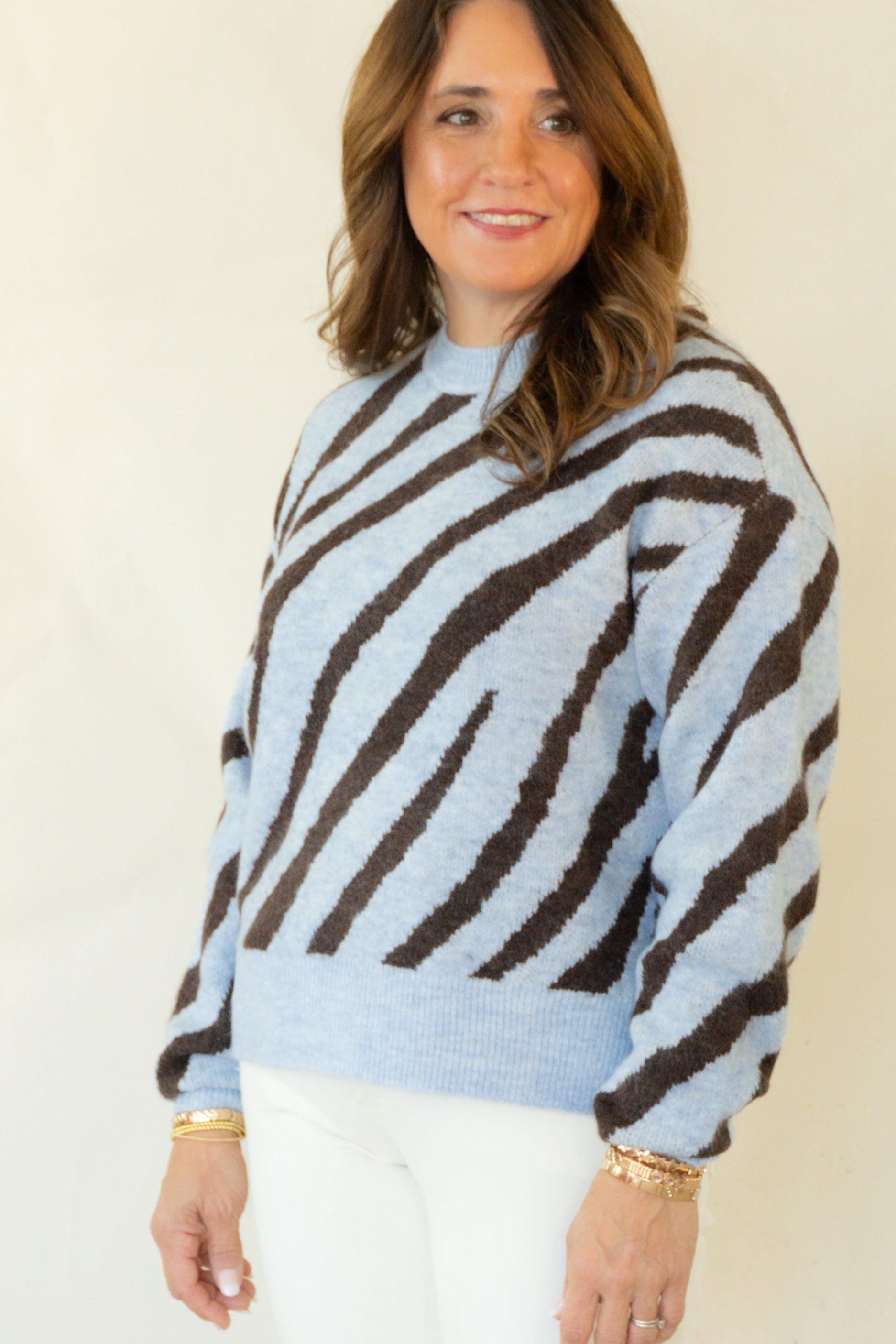A woman modeling a light blue and dark brown zebra-striped knit sweater, paired with white pants and brown shoes, standing against a plain off-white studio background.