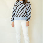 A woman modeling a light blue and dark brown zebra-striped knit sweater, paired with white pants and brown leather loafers, against a plain cream background.