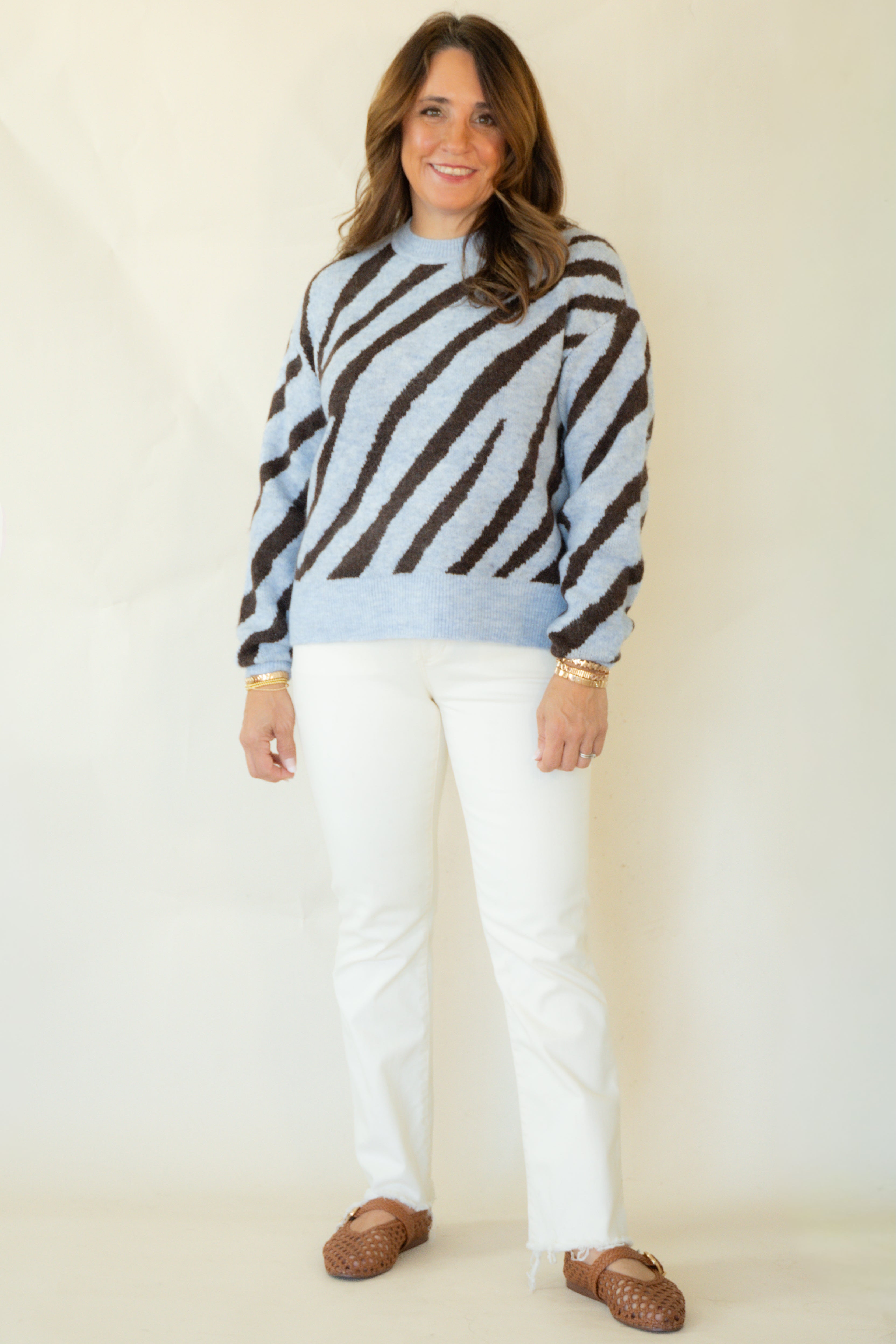A woman modeling a light blue and dark brown zebra-striped knit sweater, paired with white pants and brown leather loafers, against a plain cream background.