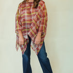 A woman modeling a rust red and olive green plaid poncho-style top with blue jeans and brown shoes, standing against a plain cream background.