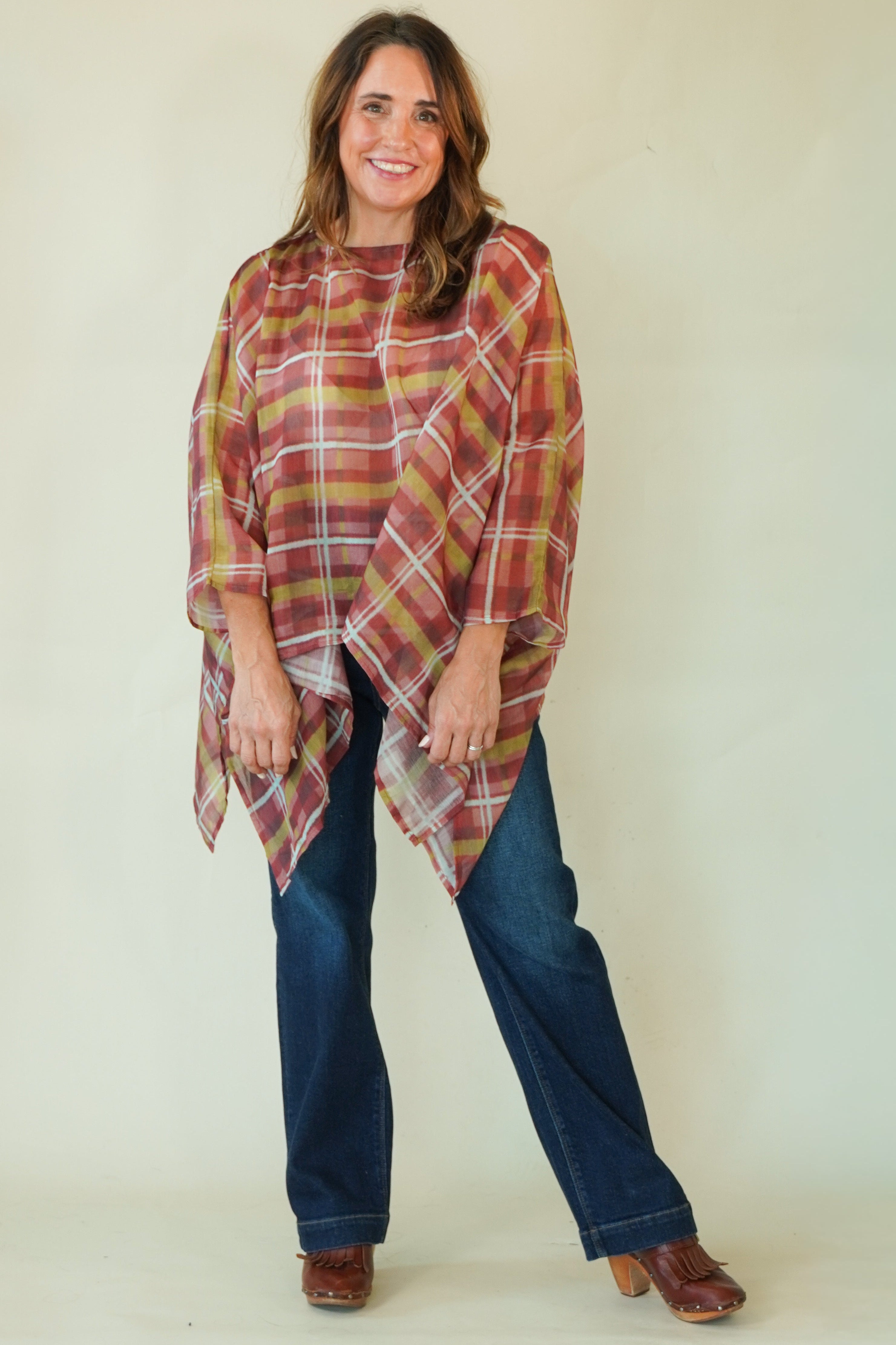 A woman modeling a rust red and olive green plaid poncho-style top with blue jeans and brown shoes, standing against a plain cream background.
