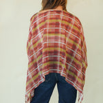 A woman viewed from behind wearing a loose, semi-sheer plaid poncho in rust red, mustard yellow, and white over dark blue jeans, set against a plain off-white background.