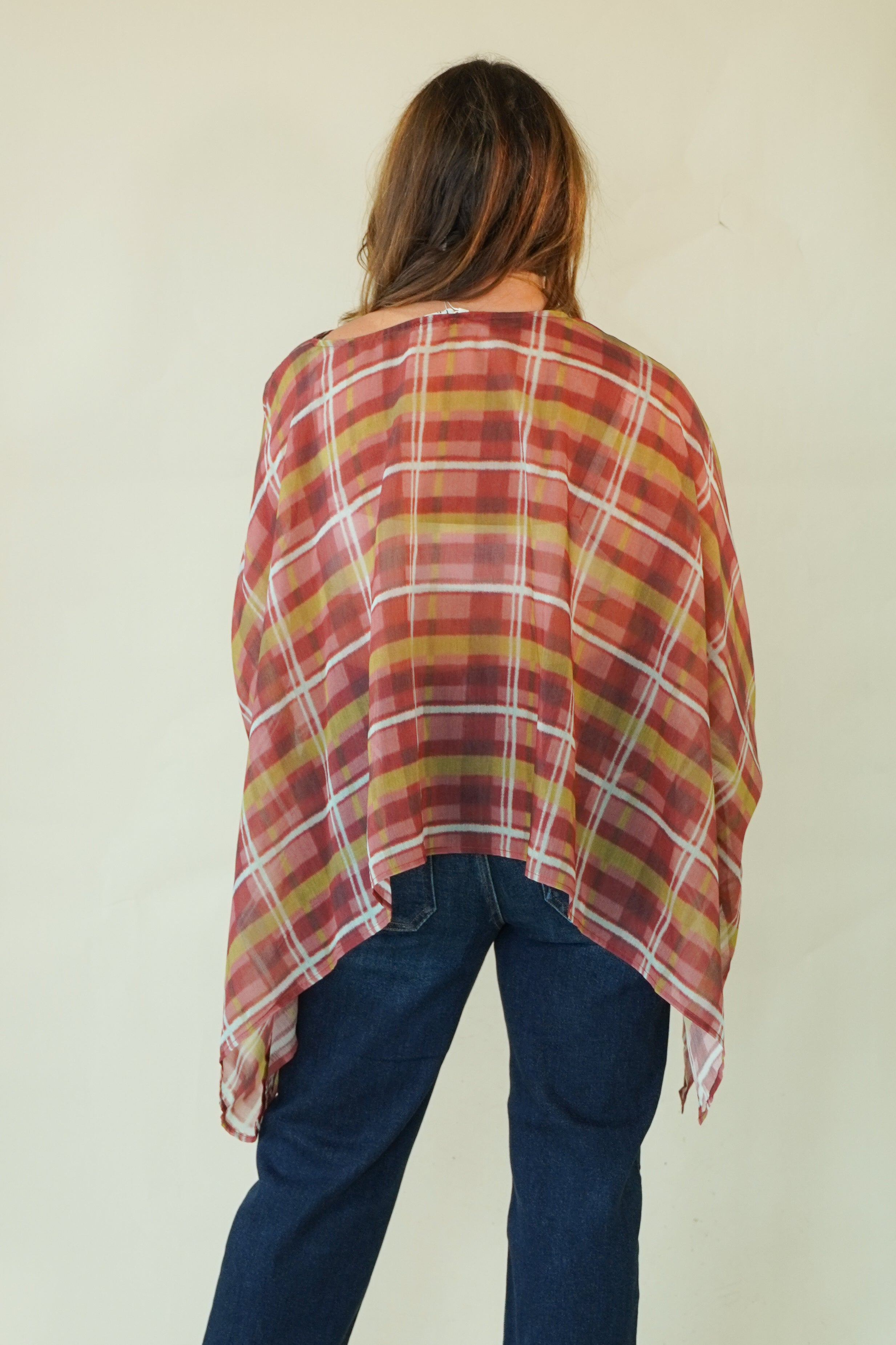 A woman viewed from behind wearing a loose, semi-sheer plaid poncho in rust red, mustard yellow, and white over dark blue jeans, set against a plain off-white background.