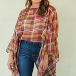 A woman modeling a rust-orange and yellow plaid poncho-style top with blue jeans and brown boots, standing against a plain light beige studio background.